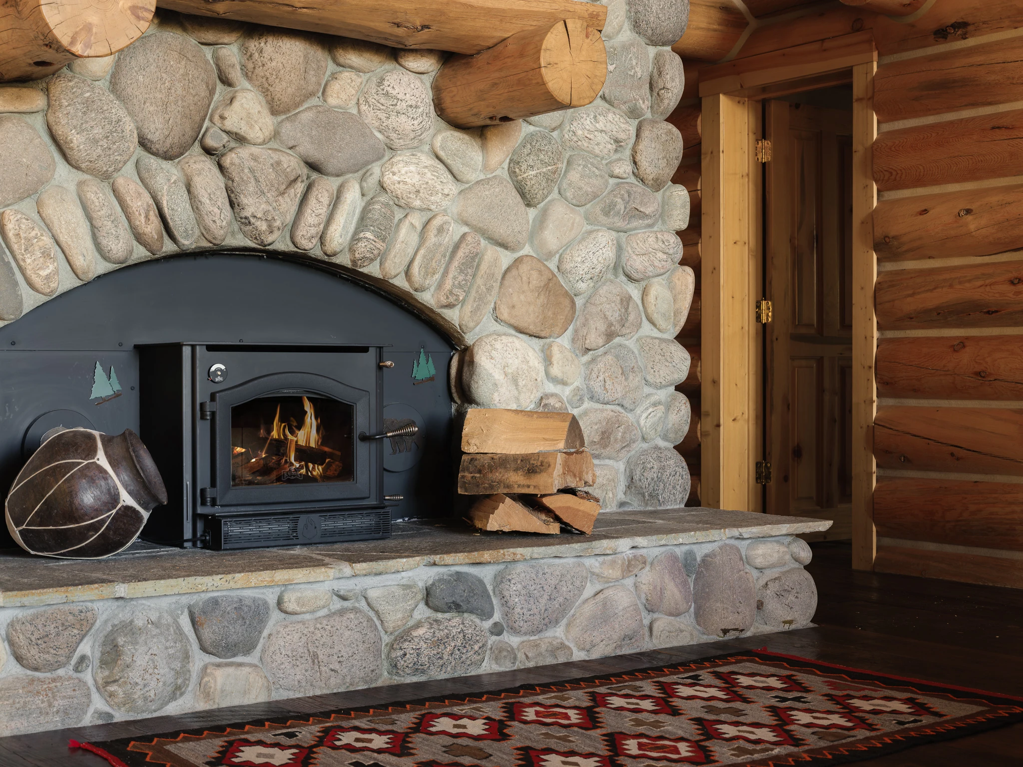 Detail of a river-rock wood-burning stove with stacked firewood — architectural photography Livingston Montana, Bear Canyon Ranch